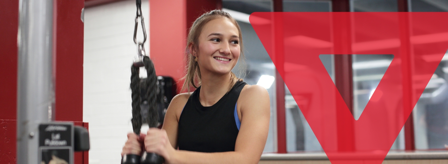 Young woman smiling while using gym equipment at the YMCA. A large transparent red YMCA logo overlays the right side of the image.