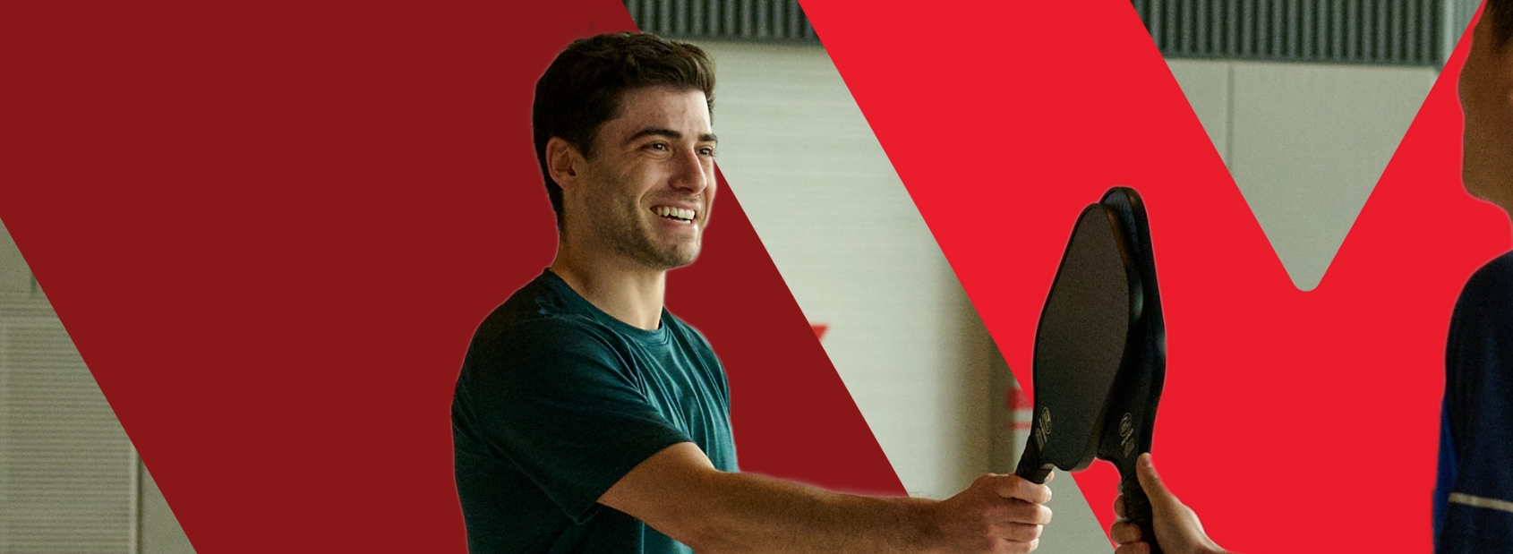 Two men on an indoor court smiling and tapping pickleball paddles in a friendly greeting, with a large stylized red YMCA logo overlaid in the background.